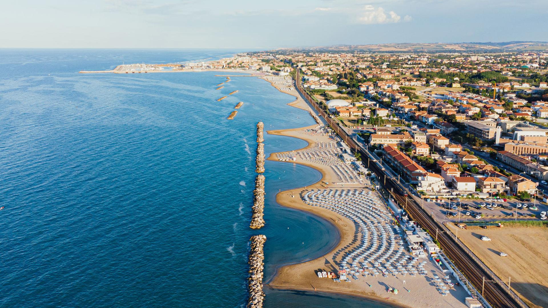 Vista aerea di una spiaggia con ombrelloni e mare calmo, città sullo sfondo.