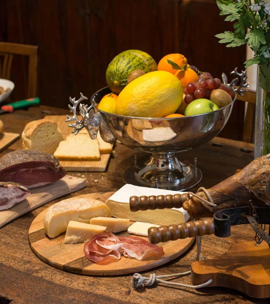Tavola imbandita con frutta, salumi, formaggi, pane e fiori.